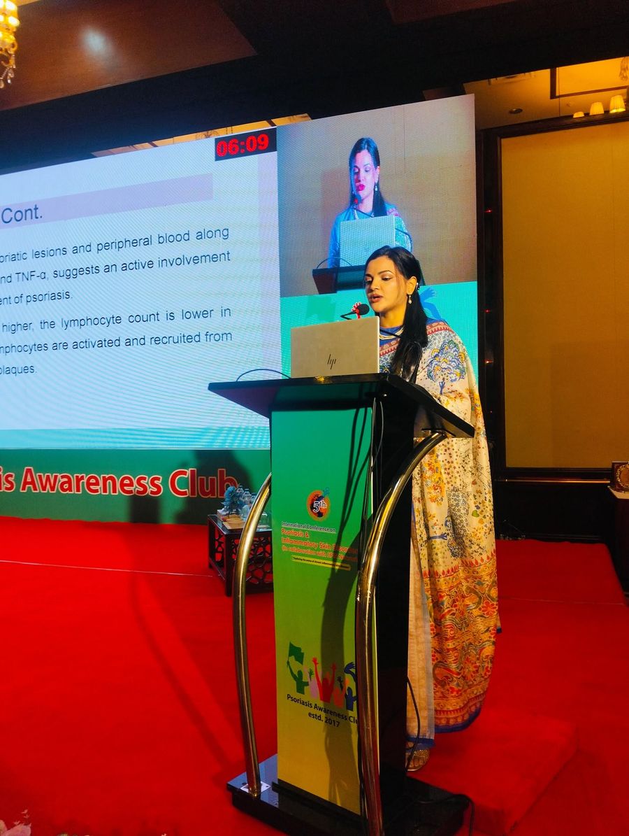 Dr. Tasmin Tahnin Siha presenting from a podium at a psoriasis conference, with a lecture slide on psoriatic lesions and a live camera feed of her on the screen behind.